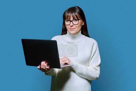 Téléchargez les photos : Belle femme d'âge moyen tenant un ordinateur portable regardant la caméra sur fond de studio bleu. Sourire femme heureuse en pull blanc à l'aide d'un ordinateur. Technologies pour les entreprises communications de travail loisirs - en image libre de droit