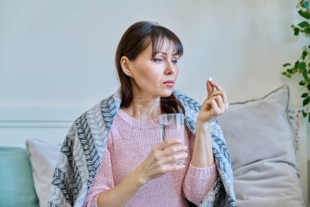 Photo for Sick mature woman taking pill with glass of water, sitting under blanket at home. Medicines, health, treatment concept - Royalty Free Image