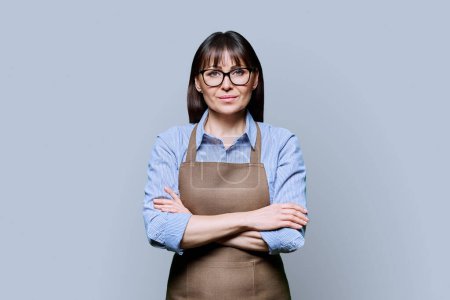 Confident serious middle-aged woman in apron on gray background. Successful mature female small business owner, service worker, entrepreneur looking at camera with crossed arms. Business work people