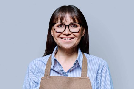Confident serious middle-aged woman in apron on grey background. Successful mature female small business owner service worker entrepreneur looking at camera. Business work people startup advertising