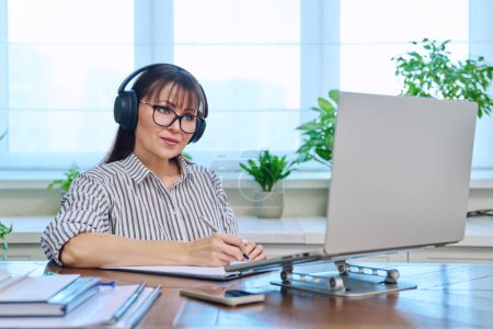 Photo for Middle-aged woman in headphones working at computer in home office, looking at web cam talking. Online meeting, remote work teaching financial legal advice interview blogging freelance, mental therapy - Royalty Free Image