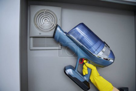 Photo for Cleaning wall-mounted hood ventilation grill in bathroom with vacuum cleaner. Housekeeping, housework, housecleaning, cleaning service concept - Royalty Free Image