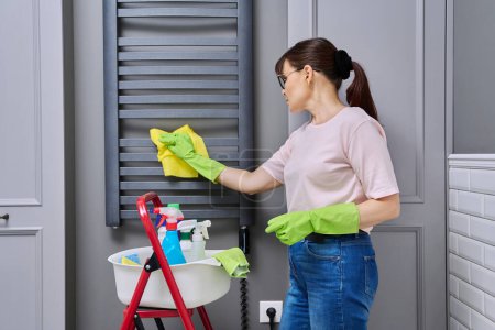 Téléchargez les photos : Femme faisant le nettoyage de la maison dans la salle de bain, nettoyer la poussière du porte-serviettes chauffant. Entretien ménager, travaux ménagers, ménage, concept de service de nettoyage - en image libre de droit