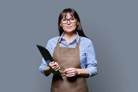 Photo for Confident smiling middle-aged woman in apron holding clipboard on grey background. Successful mature female small business owner service worker entrepreneur looking at camera. Business work people - Royalty Free Image