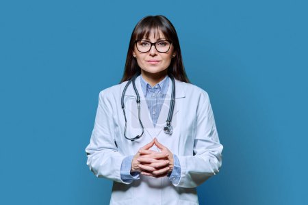 Photo for Confident friendly smiling middle-aged female doctor in white lab coat with stethoscope, looking at camera on blue studio background. Healthcare, medicine, staff, treatment, medical services concept - Royalty Free Image