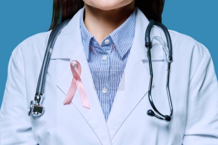 Téléchargez les photos : Gros plan femme médecin en blouse blanche avec ruban rose symbolisant la Journée internationale du cancer du sein sur fond bleu. Diagnostic de traitement du cancer du sein, médecine, santé des femmes - en image libre de droit