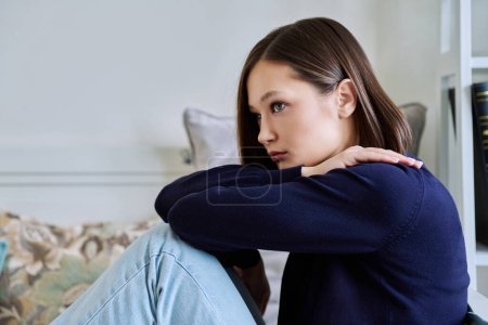 Young sad upset unhappy woman sitting on couch at home. Frustrated confused female experiencing difficulties. Mental problems, feelings, loneliness, stress, depression, difficulties, youth concept