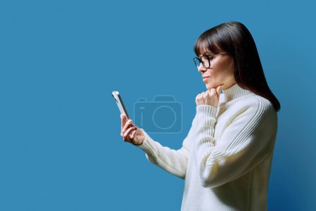 Foto de Perfil ver mujer seria de mediana edad utilizando teléfono inteligente sobre fondo azul. Mujer madura mirando el teléfono en las manos. Tecnologías aplicaciones móviles Internet trabajo negocios ocio comunicación - Imagen libre de derechos