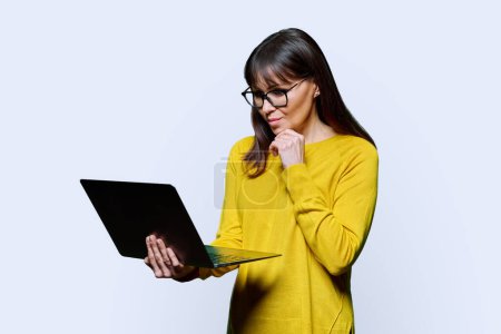 Photo for Middle-aged woman in yellow using laptop on white studio background. Serious mature female looking at computer. Technologies network for communication work business lifestyle 40s people concept - Royalty Free Image