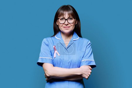 Téléchargez les photos : Portrait d'une infirmière souriante en uniforme bleu avec un ruban rose symbolisant la Journée internationale du cancer du sein sur fond bleu. Diagnostic de traitement du cancer du sein, médecine concept de santé des femmes - en image libre de droit