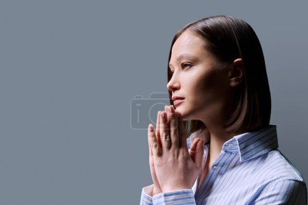 Photo for Close-up profile view of young woman with folded hands on gray background, copy space. Serious female worried praying thinking. Mental health psychology psychotherapy feelings stress depression - Royalty Free Image