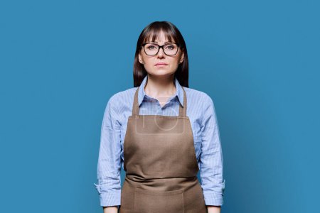 Confident serious middle-aged woman in apron on blue background. Successful mature female small business owner, service worker, entrepreneur looking at camera. Business work people startup advertising