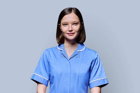 Portrait of young confident smiling female nurse looking at camera on gray studio background. Medical services, health, professional assistance, medical care concept