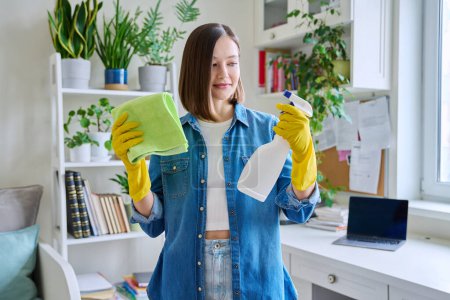 Photo for Young woman doing house cleaning, wearing gloves with cleaning detergent agent spray and rag in room interior. Routine home cleaning, cleanliness, housework, housekeeping, household, lifestyle - Royalty Free Image