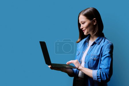 Young smiling woman using laptop computer, profile view on blue studio background, copy space for advertising text image. Internet online technology, education training business work service lifestyle