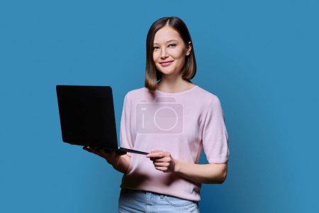 Foto de Mujer joven sonriente con ordenador portátil en fondo de estudio azul. Mujer atractiva exitosa mirando a la cámara. Negocios, trabajo, trabajo, estudio, educación, e-learning, comunicación, concepto de tecnología - Imagen libre de derechos