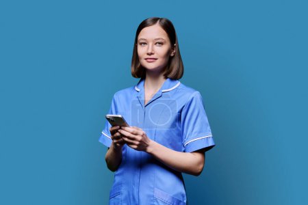Lächelnde junge Krankenschwester mit Smartphone, Blick in die Kamera auf blauem Studiohintergrund. Mobile Apps Anwendungen, Technologien in den Bereichen medizinische Dienstleistungen, Gesundheit, professionelle Hilfe, medizinische Versorgung