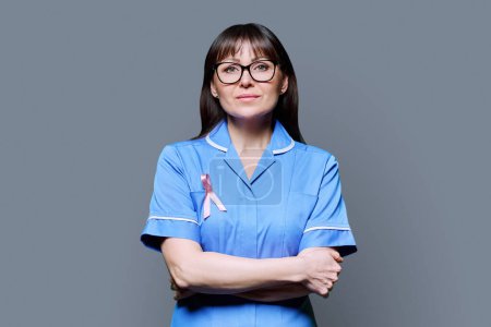 Téléchargez les photos : Portrait d'une infirmière sérieuse en uniforme bleu avec un ruban rose symbolisant la Journée internationale du cancer du sein sur fond gris. Diagnostic de traitement du cancer du sein, médecine concept de santé des femmes - en image libre de droit