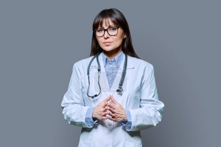 Photo for Confident serious middle-aged female doctor in white lab coat with stethoscope, looking at camera on gray studio background. Healthcare, medicine, staff, treatment, medical services concept - Royalty Free Image
