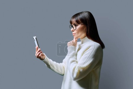 Foto de Perfil ver mujer seria de mediana edad utilizando teléfono inteligente sobre fondo gris. Mujer madura mirando el teléfono en las manos. Tecnologías aplicaciones móviles Internet trabajo negocios ocio comunicación - Imagen libre de derechos