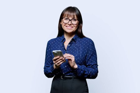 Photo for Business mature woman holding phone on white background. Smiling successful middle-aged female is texting chatting looking at camera. Mobile online internet applications for work leisure communication - Royalty Free Image