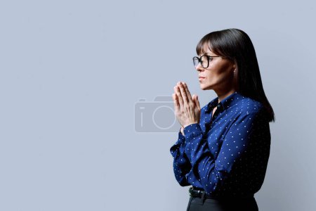 Téléchargez les photos : Femme mûre sérieuse plia les mains en prière, méditation sur fond de studio gris. Vue de profil de rêver, penser, femme d'âge moyen, espace de copie. Santé mentale, concept humain - en image libre de droit