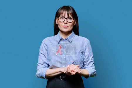 Photo for Middle-aged woman with pink ribbon on her chest symbolizing International Breast Cancer Day, on blue studio background. Solidarity, patient support, health care, treatment, medicine, world cancer day - Royalty Free Image