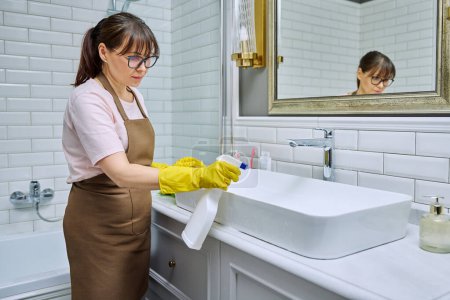 Photo for Woman in apron with detergent washcloth cleaning in bathroom, washing sink washbasin. Housewife cleaning house, cleaner service worker at workplace. Home hygiene housecleaning housekeeping housework - Royalty Free Image