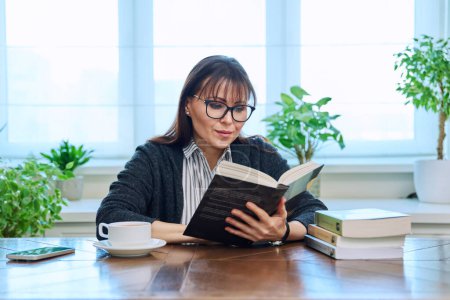 Photo for Middle-aged woman reading books, sitting at the table at home. Literary hobby, leisure, relaxation, people concept - Royalty Free Image