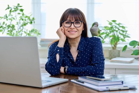 Foto de Mujer de mediana edad sentada en el lugar de trabajo con computadora en la oficina en casa, sosteniendo a su mascota con loro cuáquero verde en su hombro. Trabajo, estilo de vida, mascotas, aves tropicales - Imagen libre de derechos