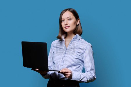 Téléchargez les photos : Sérieuse jeune femme d'affaires avec ordinateur portable sur fond bleu. Femme confiante qui tient l'ordinateur dans les mains en regardant la caméra. Affaires, travail, formation, éducation, technologies numériques modernes - en image libre de droit