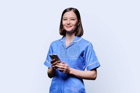 Lächelnde junge Krankenschwester mit Smartphone, Blick in die Kamera auf weißem Studiohintergrund. Mobile Apps Anwendungen, Technologien in den Bereichen medizinische Dienstleistungen, Gesundheit, professionelle Hilfe, medizinische Versorgung