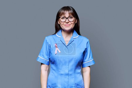 Foto de Retrato de enfermera sonriente en uniforme azul con cinta rosa que simboliza el Día Internacional del Cáncer de Mama sobre fondo gris. Tratamiento diagnóstico de cáncer de mama, medicina concepto de salud de las mujeres - Imagen libre de derechos