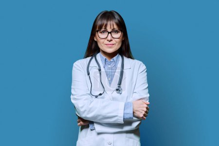 Photo for Confident friendly middle-aged female doctor in white lab coat with stethoscope, looking at camera with crossed arms on blue studio background. Healthcare, medicine, staff, treatment, medical services - Royalty Free Image
