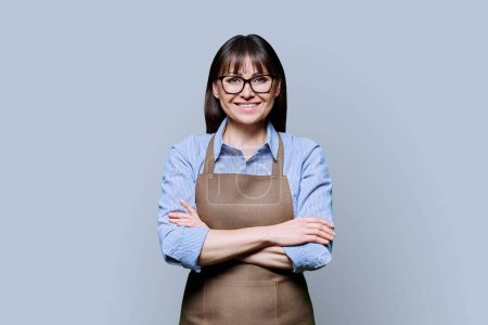 Confident smiling middle-aged woman in apron on gray background. Successful mature female small business owner, service worker, entrepreneur looking at camera with crossed arms. Business work people