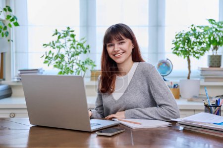 Photo for Young female college student studying at home at desk using computer laptop, writing in notebook, smiling looking at camera. E-learning, education, technology, knowledge, youth concept - Royalty Free Image