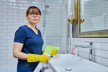 Photo for Serious woman with detergent spray, rag cleaning in bathroom, washing sink washbasin. Routine house cleaning, home hygiene, housecleaning service, housekeeping, housework concept - Royalty Free Image