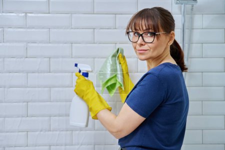 Photo for Woman with detergent spray, rag cleaning in bathroom, washing white wall tiles, smiling female looking at camera. Routine house cleaning, home hygiene, housecleaning service, housekeeping, housework - Royalty Free Image