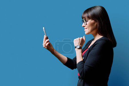 Photo for Profile view middle aged smiling woman using smartphone on blue background. Mature female looking at phone in hands. Technologies mobile apps applications internet work business leisure communication - Royalty Free Image