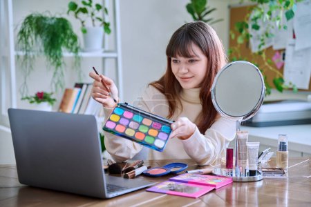 Photo for Young female showing cosmetics to laptop webcam, sitting at table at home. Beauty blog vlog, online course, training webinar on professional use of decorative cosmetics - Royalty Free Image