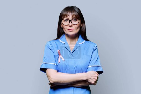 Téléchargez les photos : Portrait d'une infirmière sérieuse en uniforme bleu avec un ruban rose symbolisant la Journée internationale du cancer du sein sur fond gris. Diagnostic de traitement du cancer du sein, médecine concept de santé des femmes - en image libre de droit