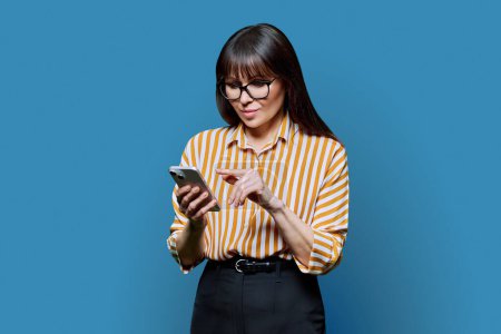 Photo for Serious business mature woman using smartphone on blue background. Middle-aged confident successful female holding phone texting reading looking. Mobile internet online technologies apps applications - Royalty Free Image