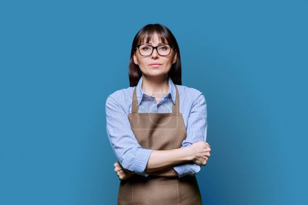 Confident serious middle-aged woman in apron on blue background. Successful mature female small business owner, service worker, entrepreneur looking at camera with crossed arms. Business work people