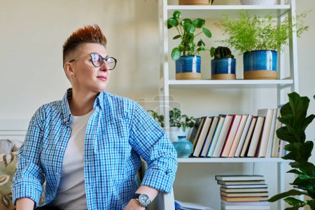 Photo for Portrait of middle-aged woman with red hair wearing glasses in casual clothes sitting on couch at home looking out window, copy space - Royalty Free Image