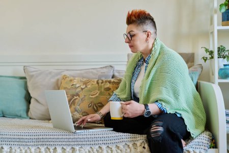 Mature woman with warm blanket holding cup of hot drink looking at laptop screen sitting on couch at home. Home coziness comfort, cold seasons, lifestyle, technology for leisure work communication