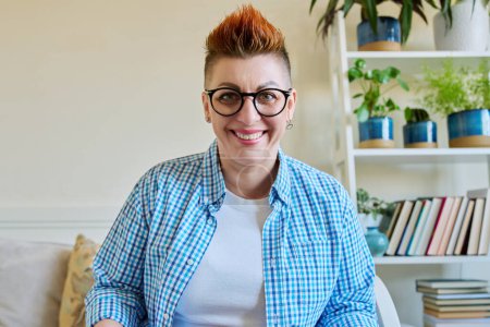 Téléchargez les photos : Portrait de femme heureuse souriante d'âge moyen dans des lunettes avec coupe de cheveux rouge regardant la caméra à l'intérieur de la maison - en image libre de droit