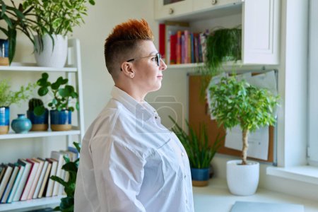 Photo for Profile portrait of serious middle-aged woman in glasses with red haircut looking at window in home interior, copy space. Mature people, lifestyle, health, life concept - Royalty Free Image