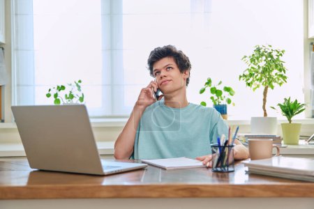 Photo for Young guy talking on the phone, sitting at his desk at home. - Royalty Free Image