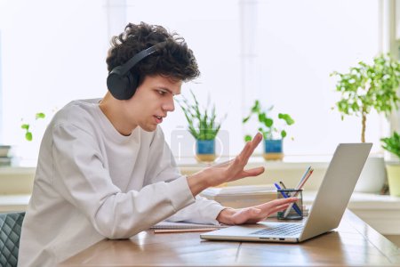 Young male student in headphones sitting at home at desk looking talking in web camera of computer laptop. Guy 19-20 years old studying remotely, video conference call, e-learning technology education