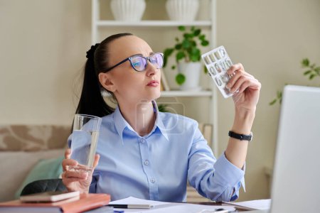 Serious tired sad woman at workplace holding blister with capsules glass water. Female taking painkiller vitamin nutritional supplement antiviral drug antioxidant antidepressant. Pharmacy health care